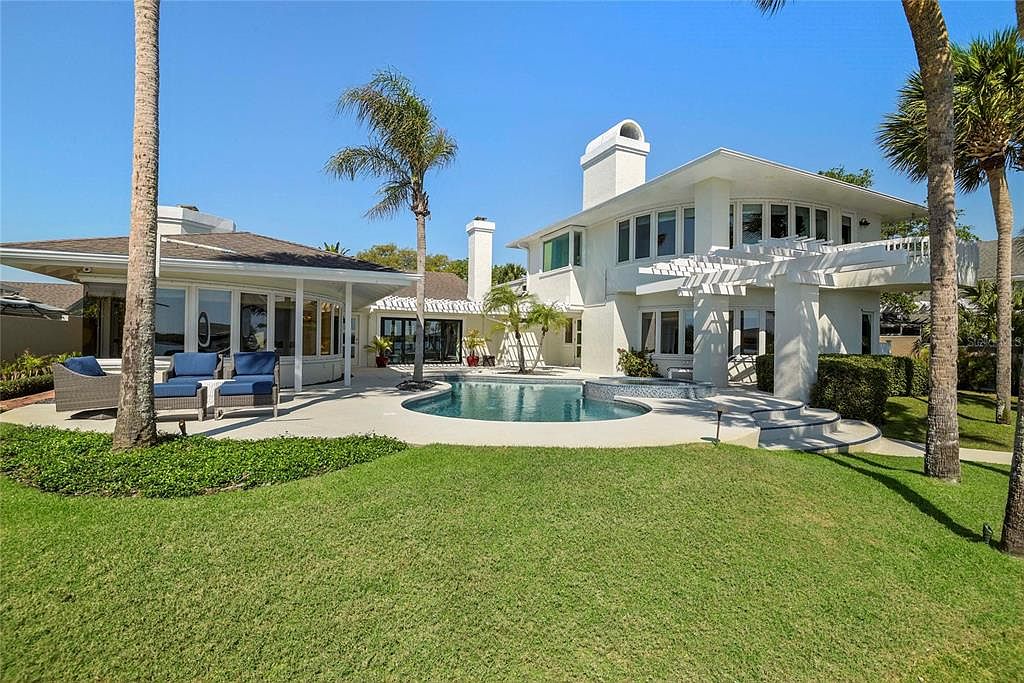Rear elevation of 1215 Commodore Drive seen across the lawn, the two-story stucco residence with a sloped roof, second-floor pergola railing, and the freeform pool with attached spa in the foreground