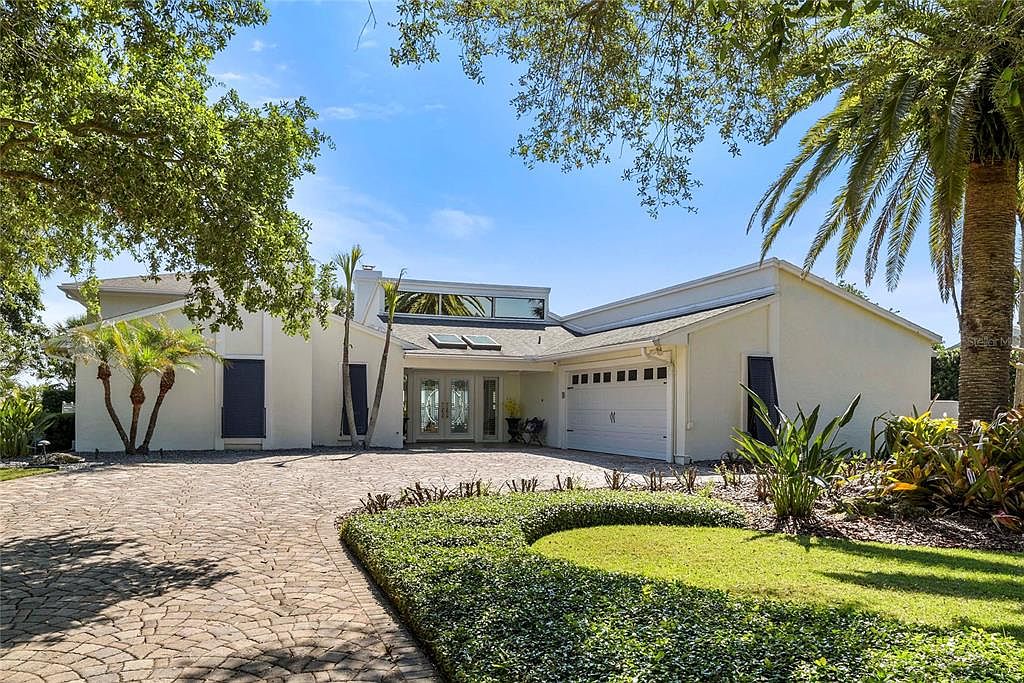 Front elevation of 1215 Commodore Drive, a one-and-two-story custom residence with white stucco, a brick paver drive court, attached garage, and tall palm to the right