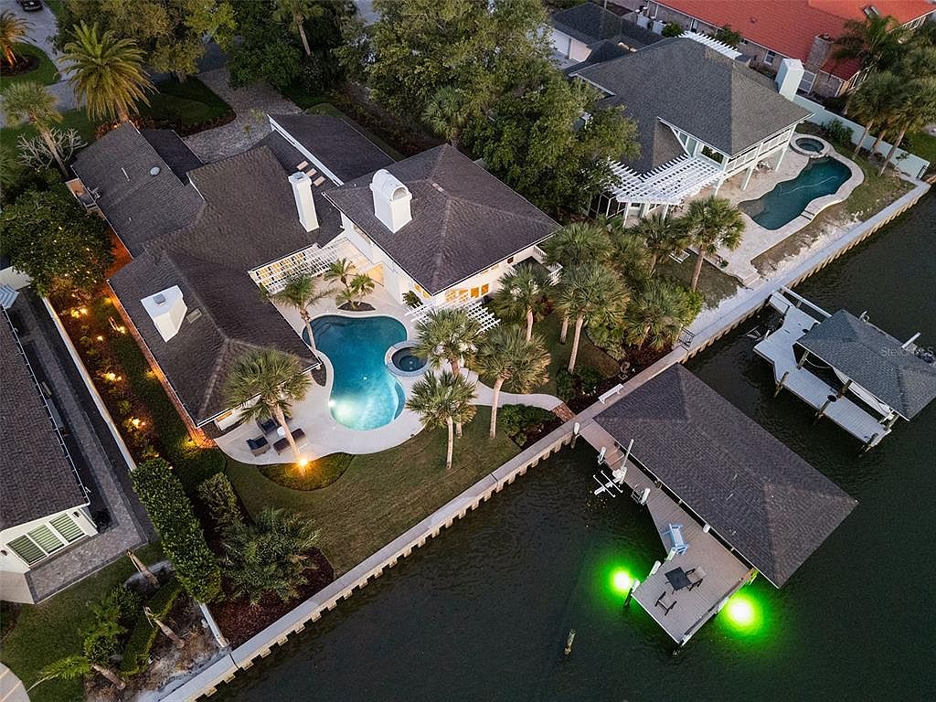 A second look at dusk over 1215 Commodore Drive from a slightly altered aerial angle, the covered boat house lit from beneath in green, jet-ski lift to its right, the dock's full footprint visible against the Indian River